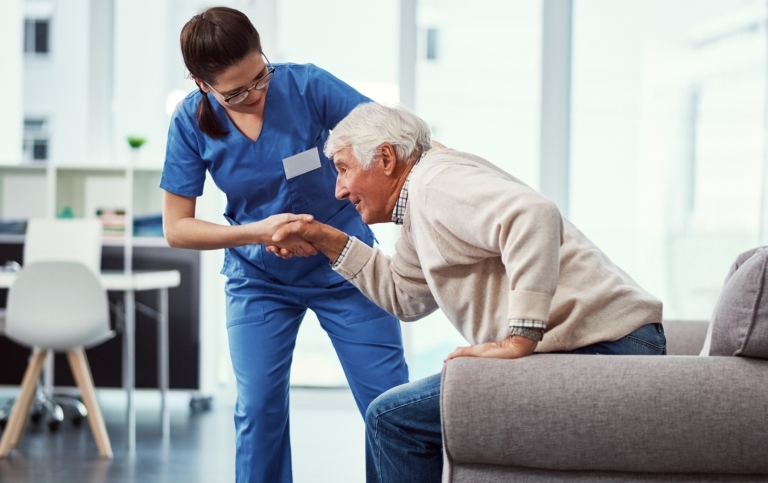 Kvinnlig vårdare hjälper en äldre man att resa sig ur en soffa